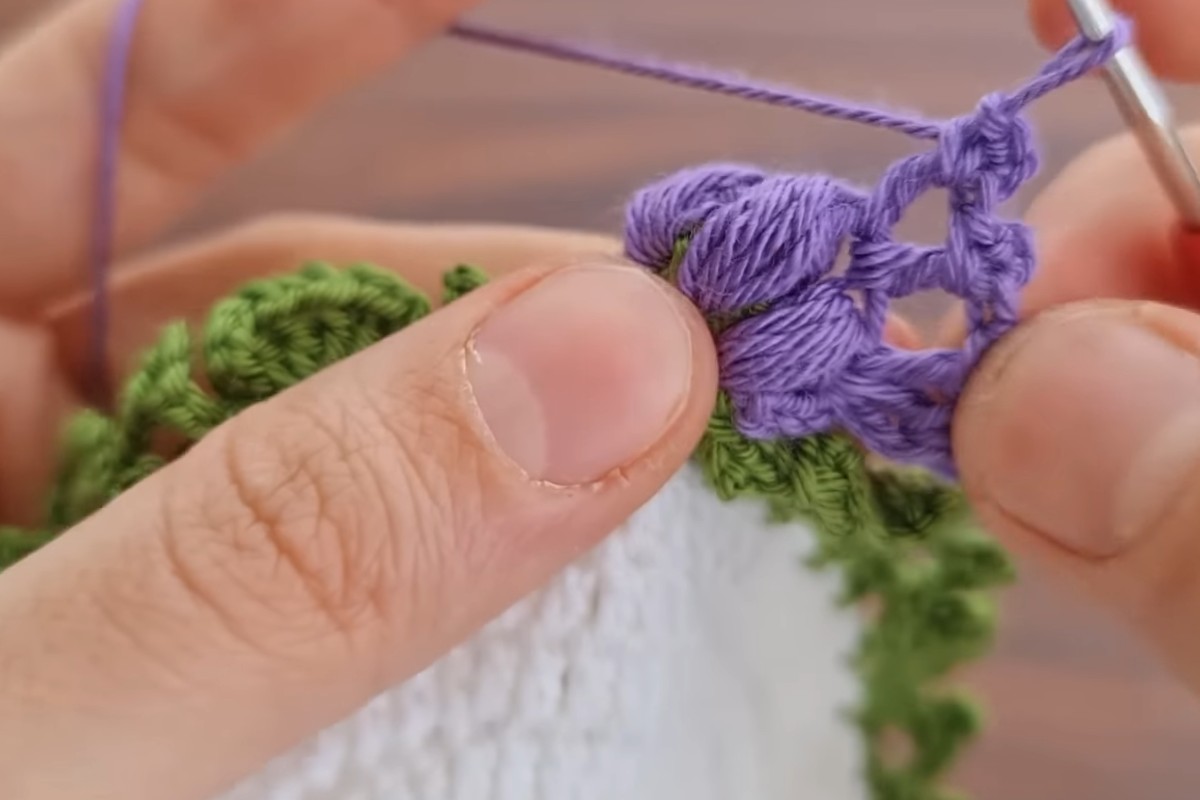Working a purple puff stitch flower on the edge of the crochet flower coaster