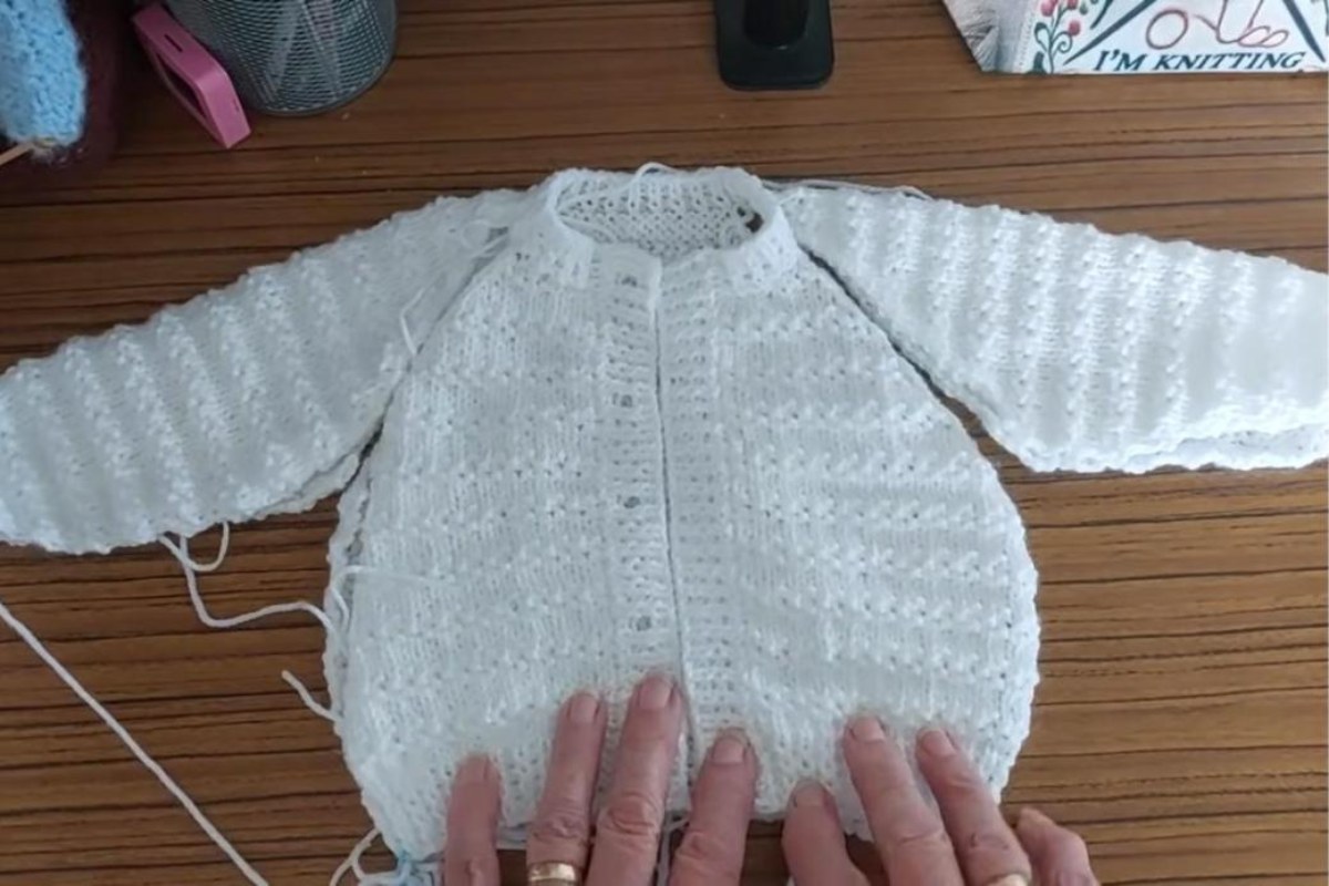 Knitted white baby cardigan laid flat with neckband and button bands done, ready for sewing the raglan and side seams